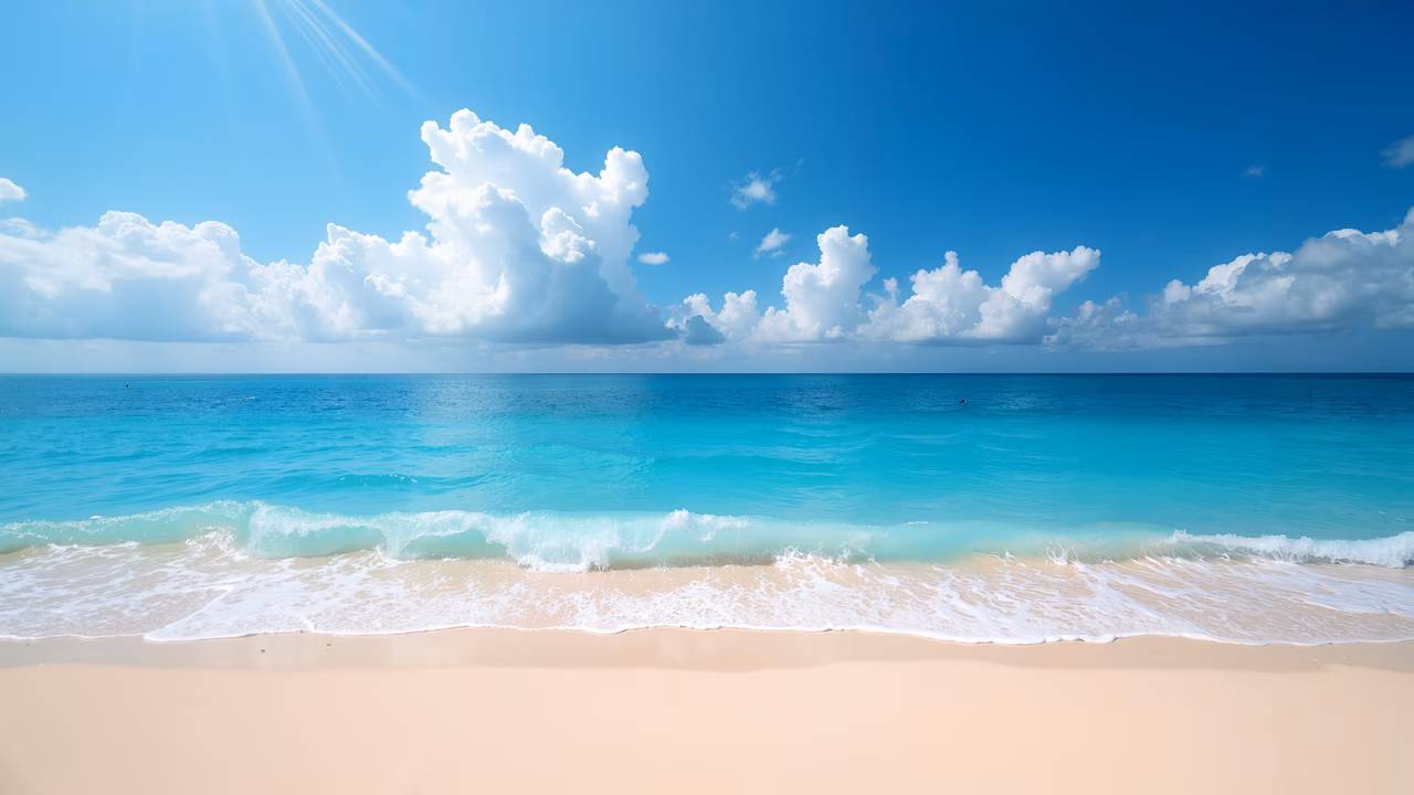 A peaceful beach with clear turquoise water and soft sand under a bright blue sky with white clouds.