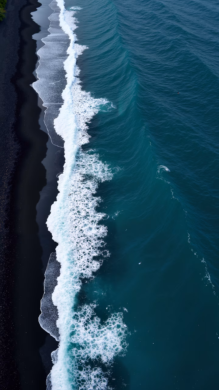 Aerial view of ocean waves meeting a black sand beach, contrasting dark sand with vibrant turquoise water, creating a tranquil natural scene.