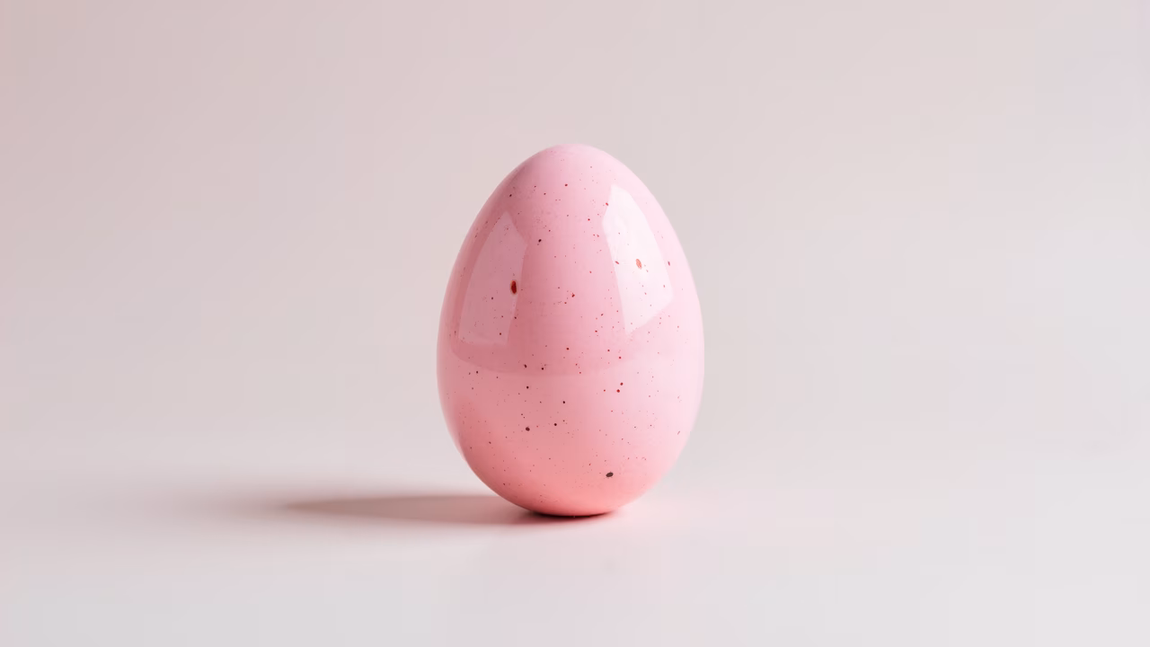Minimalist still life featuring a glossy pink speckled egg on a white background.