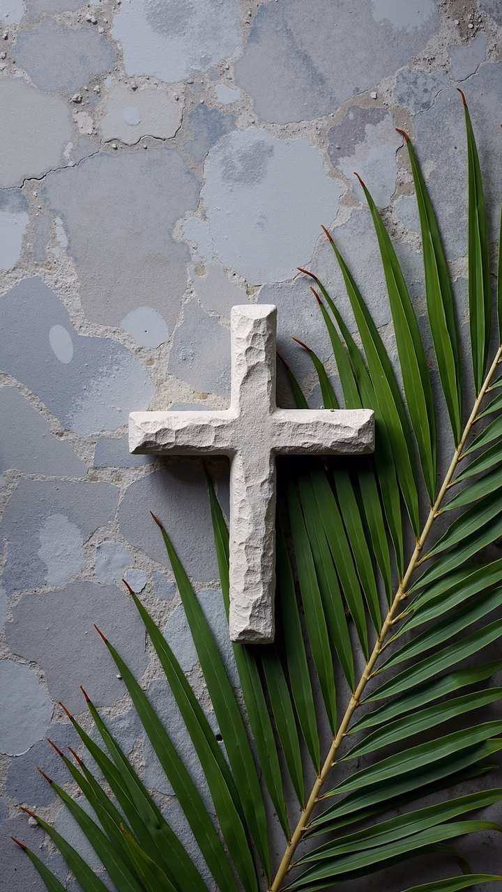 Weathered stone cross on a textured concrete wall with vibrant green palm fronds.