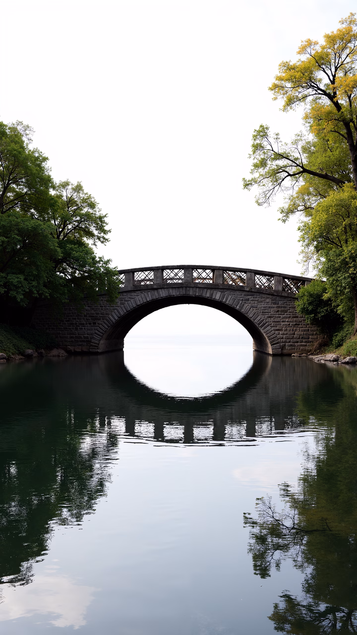 A stone bridge reflecting in a serene lake, surrounded by lush greenery.