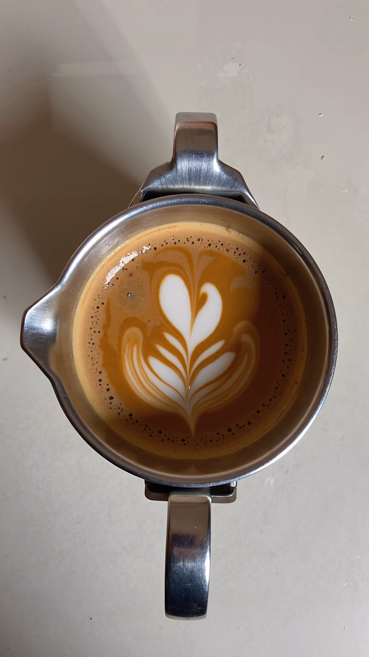 Close-up of latte art heart in a metallic pitcher, showcasing detailed coffee art against a neutral background.