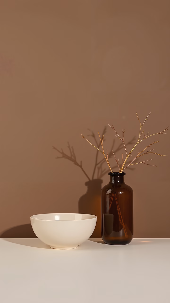 A minimalist still life with a white bowl and a dark brown vase holding dried branches, set against a muted beige backdrop.
