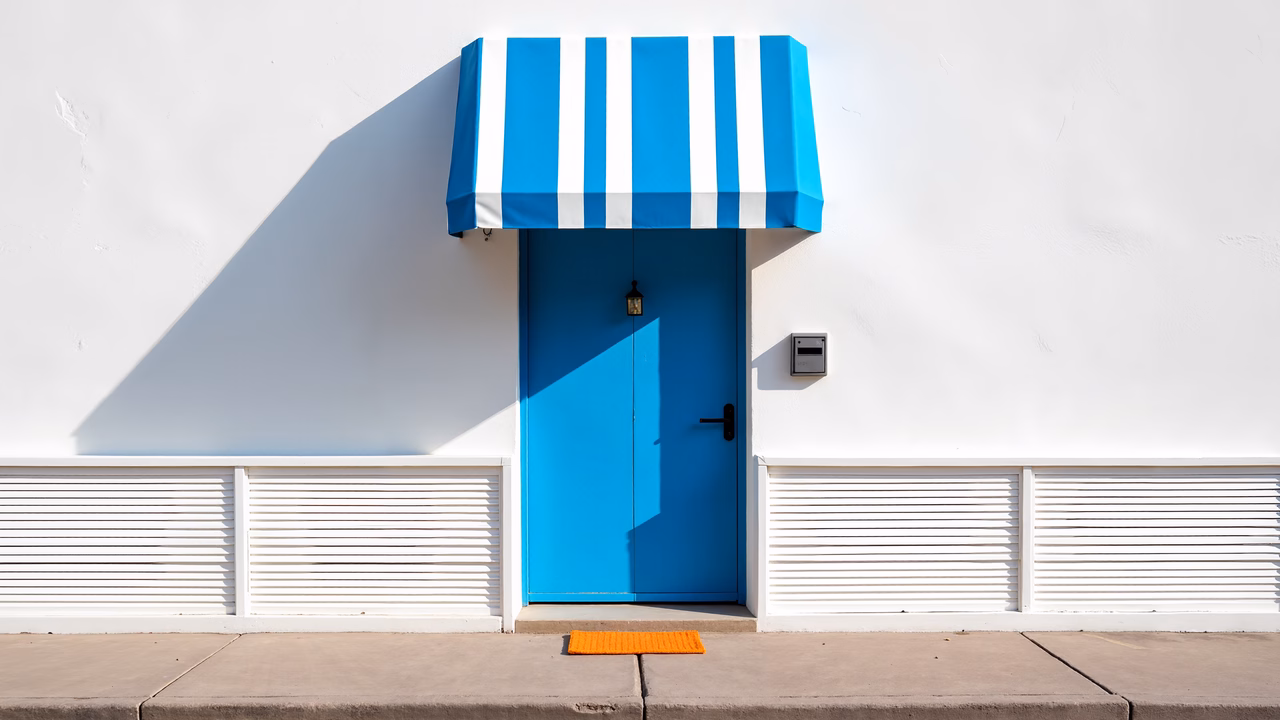 Bright blue door with striped awning on a white wall, showcasing architectural simplicity and vibrant colors.